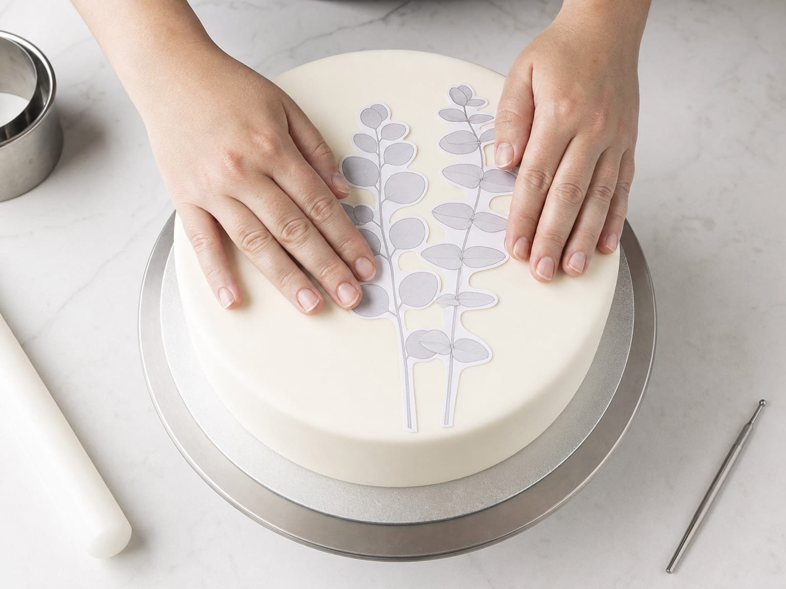 Hands laying a paper template onto a smooth fondant cake surface with a scriber tool ready