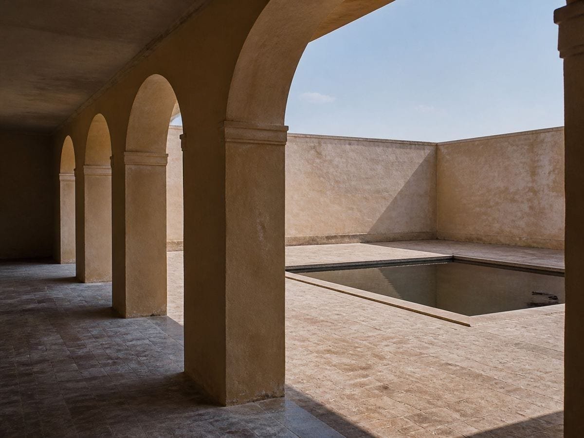 A shaded plastered courtyard arcade looking out to a still rectangular pool in sunlight