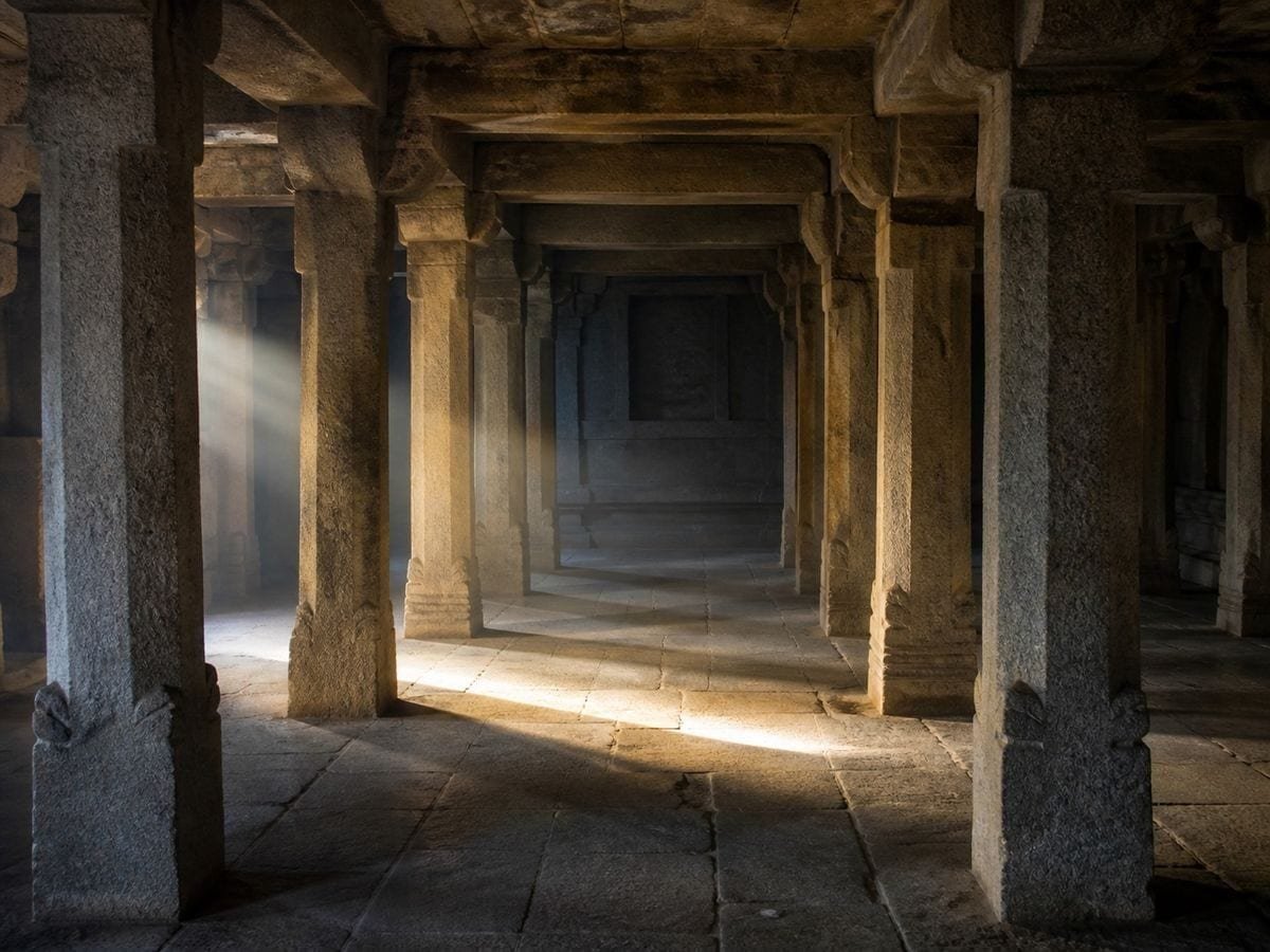 A stone temple hall of plain columns at dawn with light falling across worn flags
