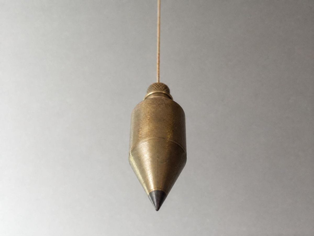 A brass plumb bob hanging still on a linen line, its point aimed toward the camera